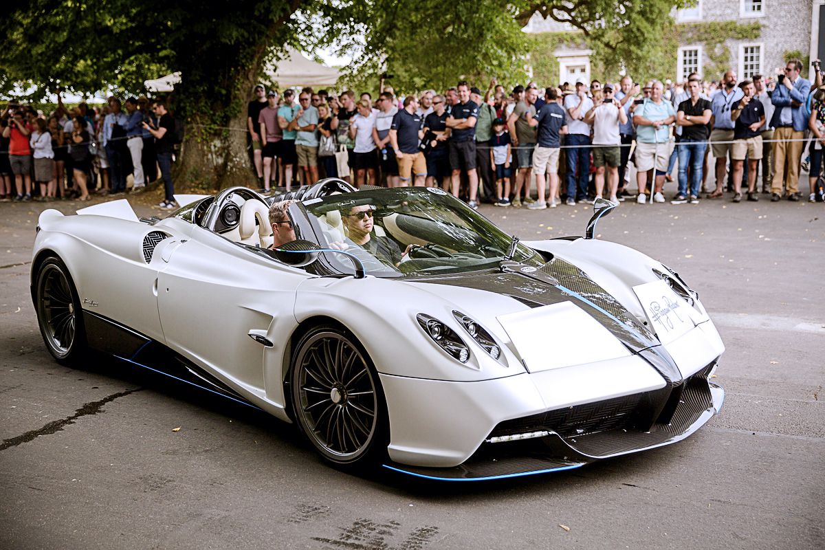 A white/grey Pagani Huayra with blue highlights facing right in front of a crowd of spectators.